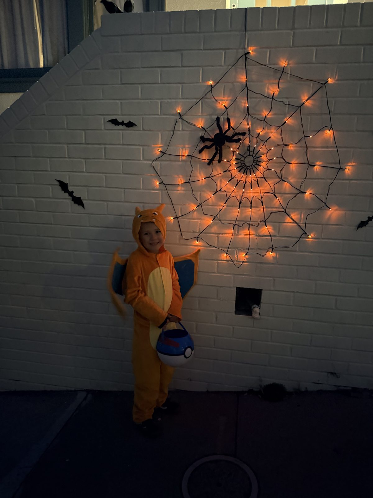 A young trick-or-treater in a dragon costume stands by a glowing orange spider web on a Somerset wall